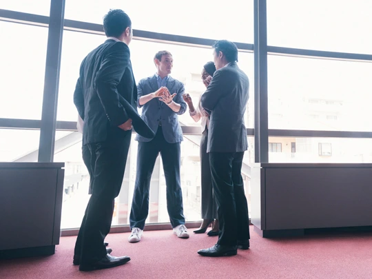 A group of investors in suits listening to a professional in front of a large window. - For your Legacy